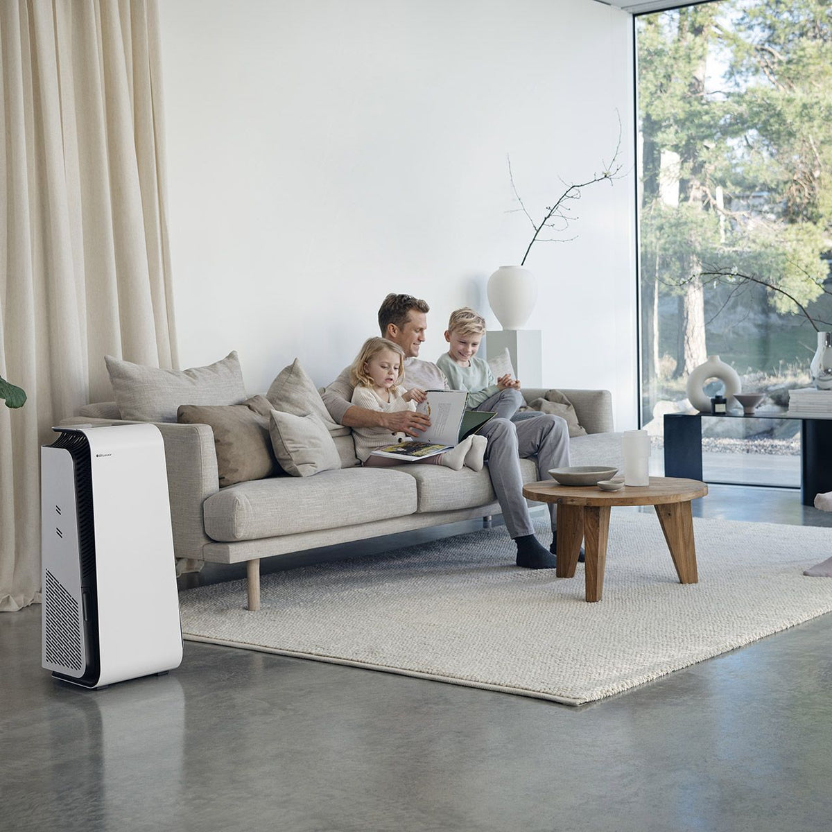 Blueair HealthProtect 7440i Luftreiniger in Weiß neben einem Sofa, Vater mit zwei Kindern im Wohnzimmer